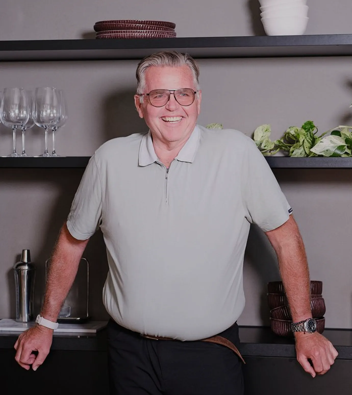 Smiling older man wearing glasses and light polo shirt leaning on a kitchen counter.