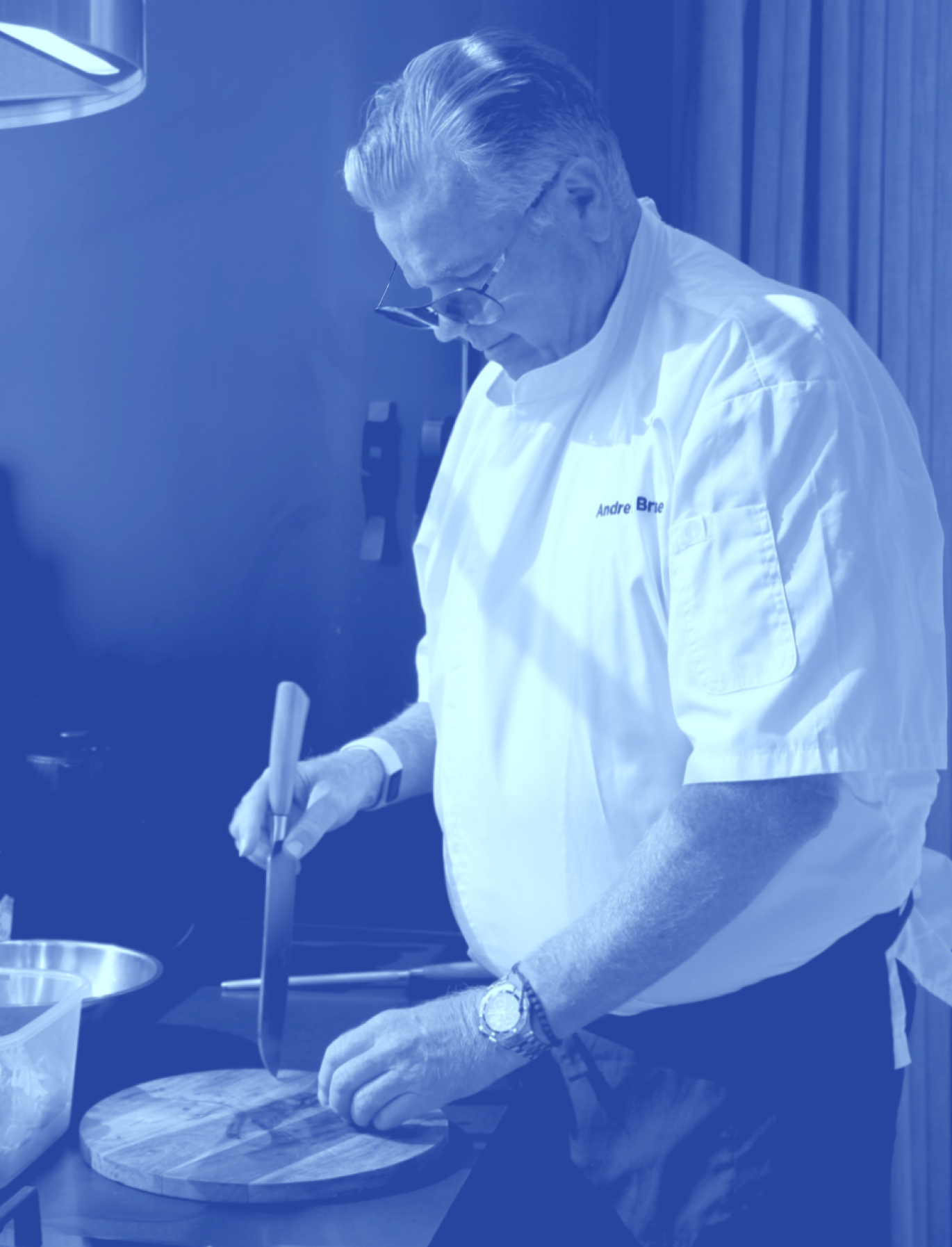 Chef in white coat slicing food on a round wooden cutting board in a kitchen.
