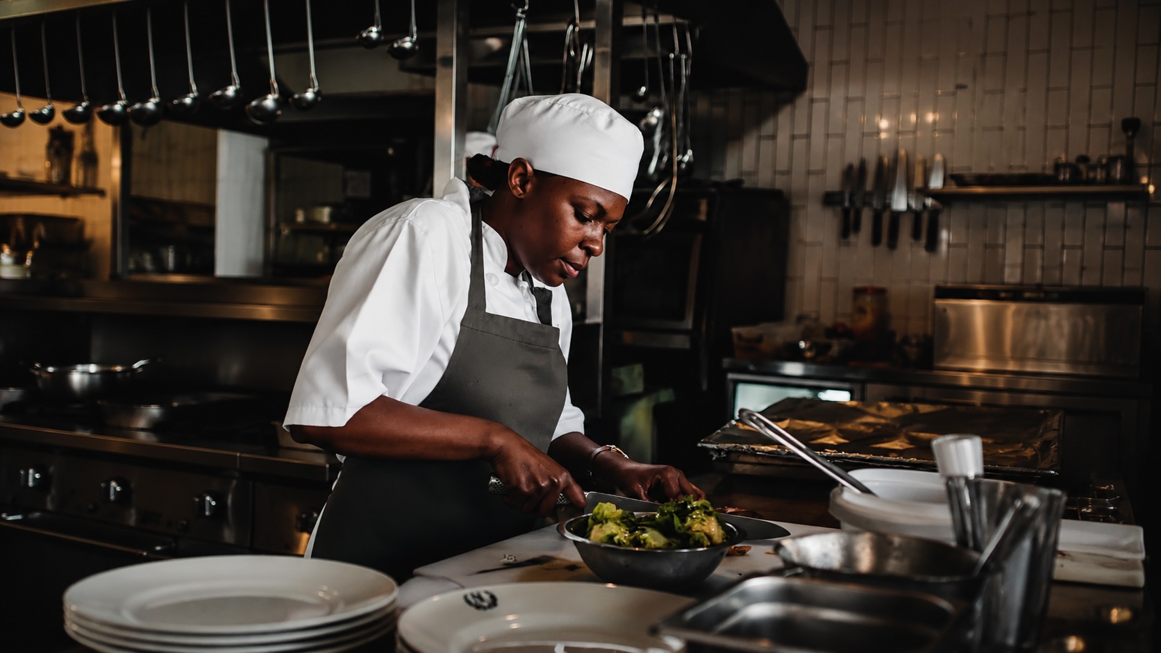 Chef in white hat and apron preparing salad in a professional kitchen with utensils and plates.