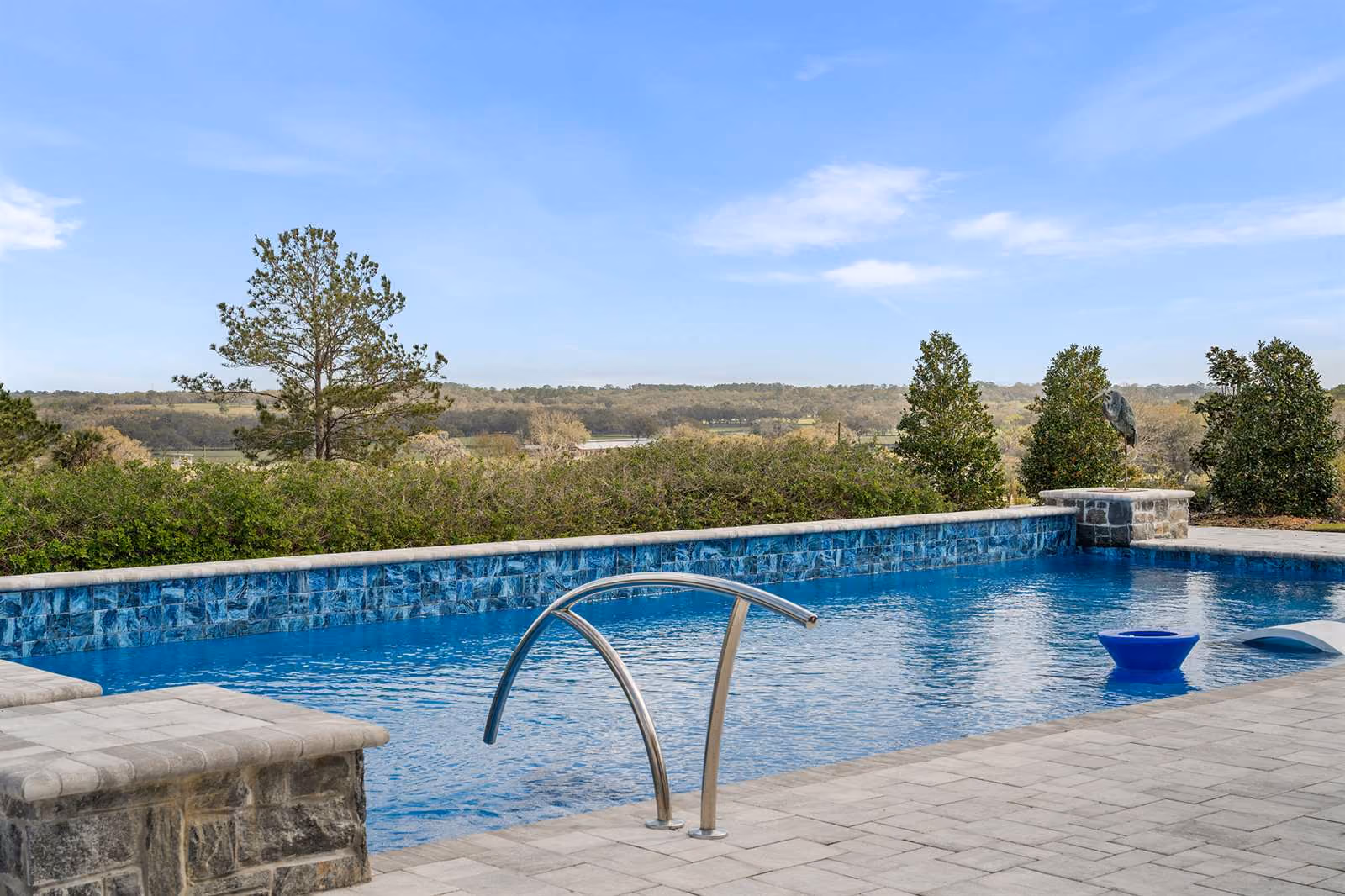 Infinity-edge pool with blue glass tile and a stainless handrail overlooking open countryside