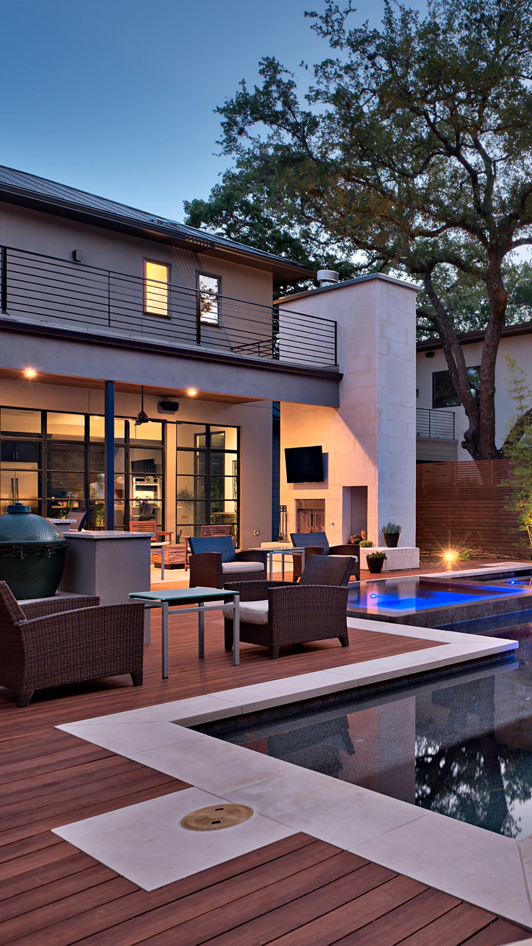 Backyard pool and wood deck at dusk with a lit patio and outdoor kitchen under live oaks