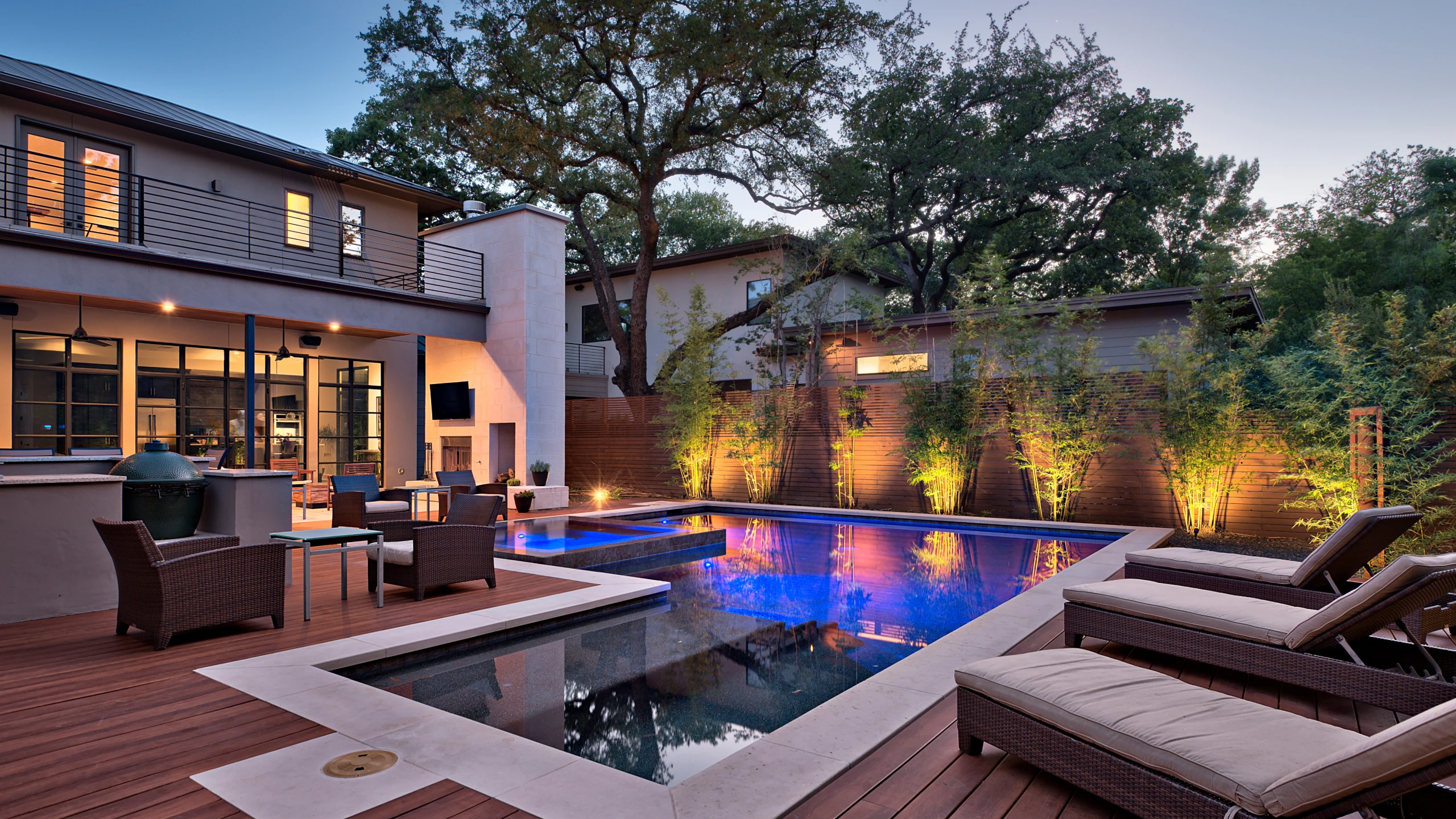 Backyard pool and wood deck at dusk with a lit patio and outdoor kitchen under live oaks