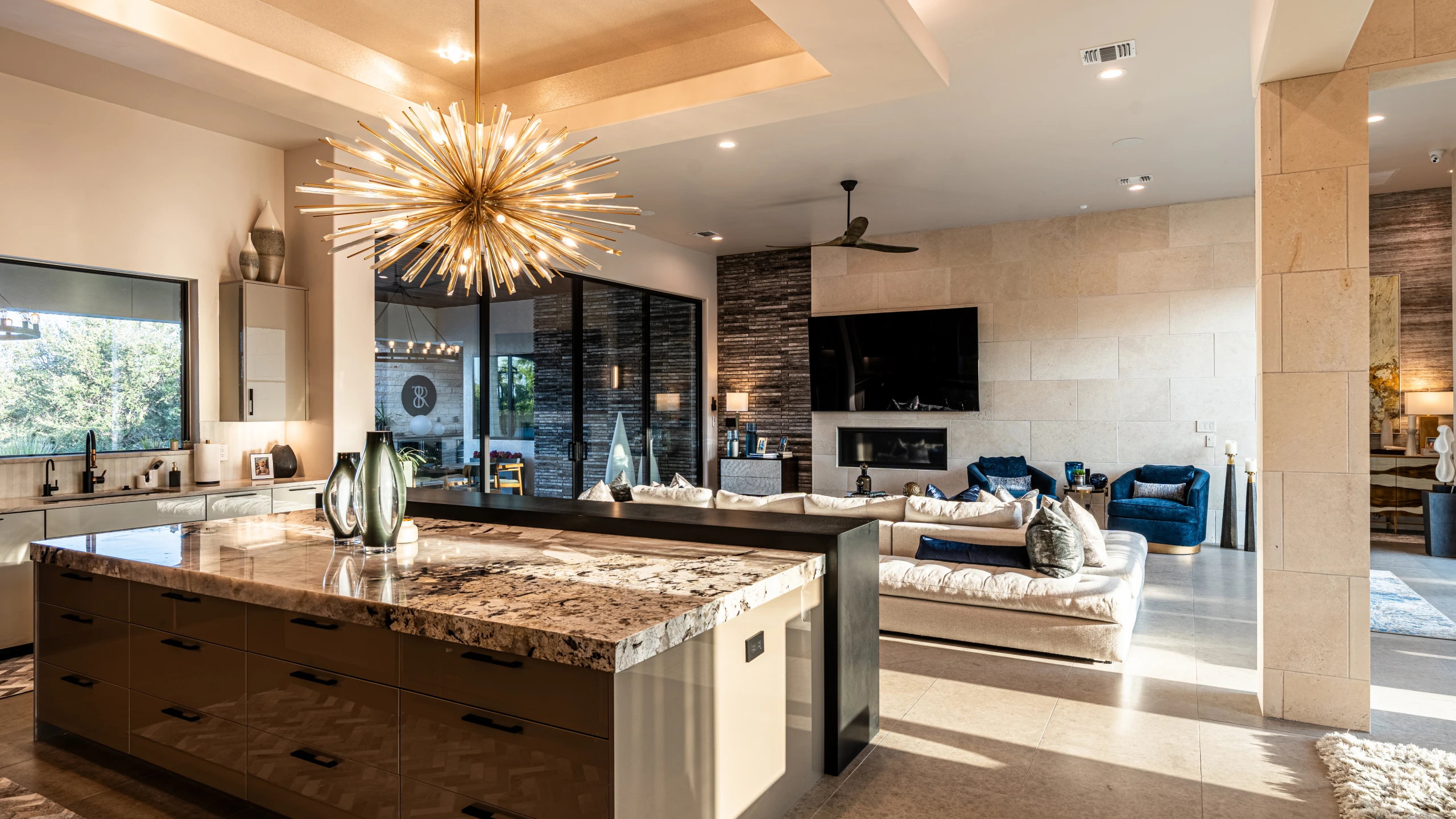 Kitchen with a granite island and starburst chandelier opening to a living room with a stone fireplace wall