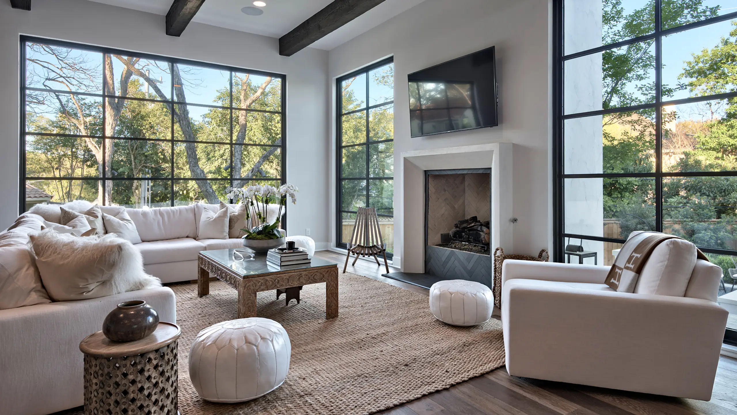 Living room with floor-to-ceiling black steel windows, a white fireplace and beamed ceiling looking out to oak trees