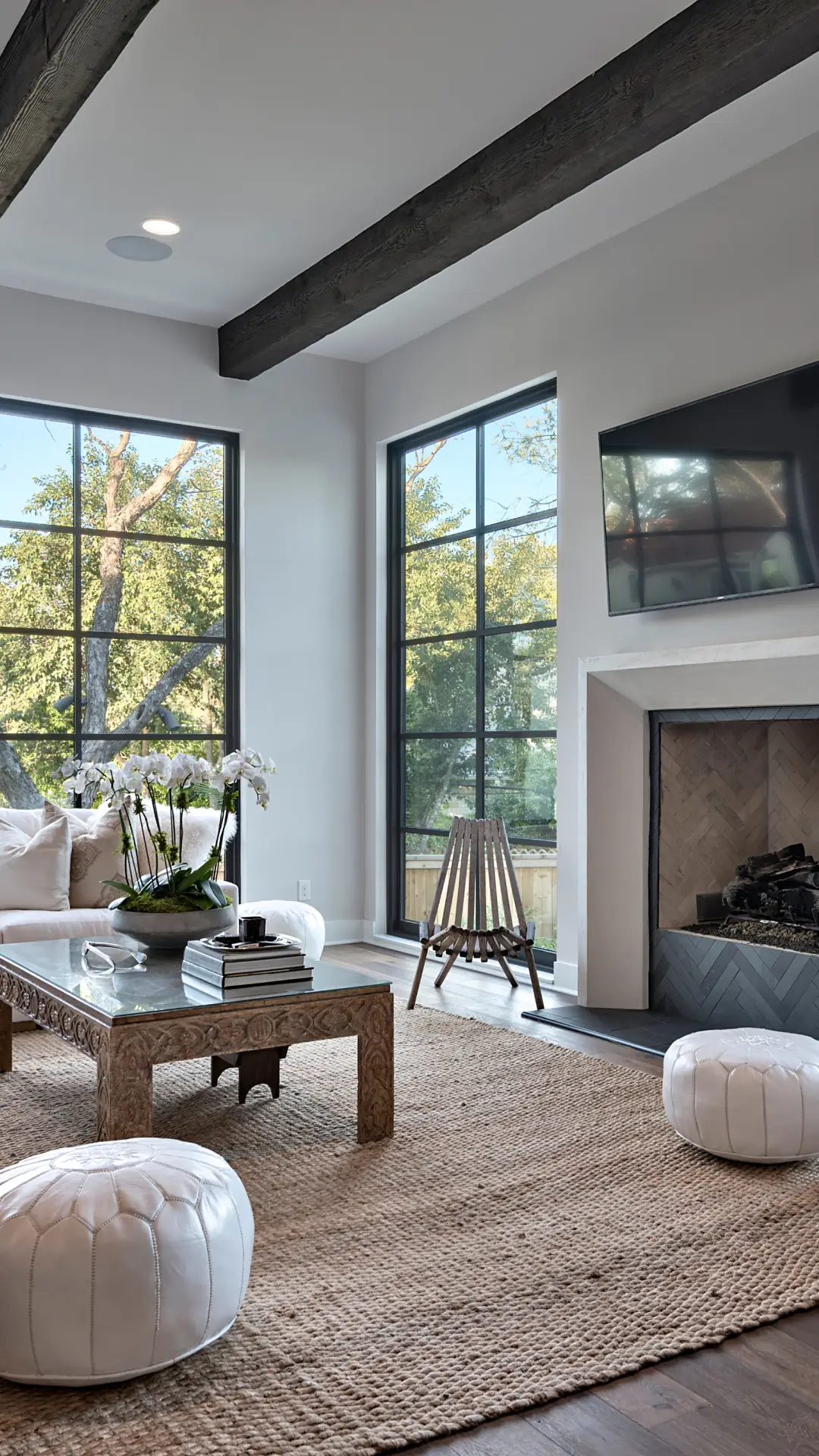 Living room with floor-to-ceiling black steel windows, a white fireplace and beamed ceiling looking out to oak trees