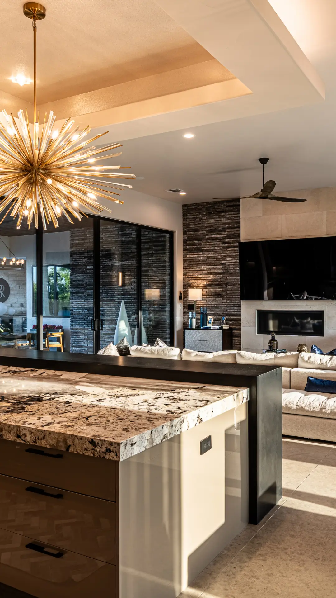 Kitchen with a granite island and starburst chandelier opening to a living room with a stone fireplace wall