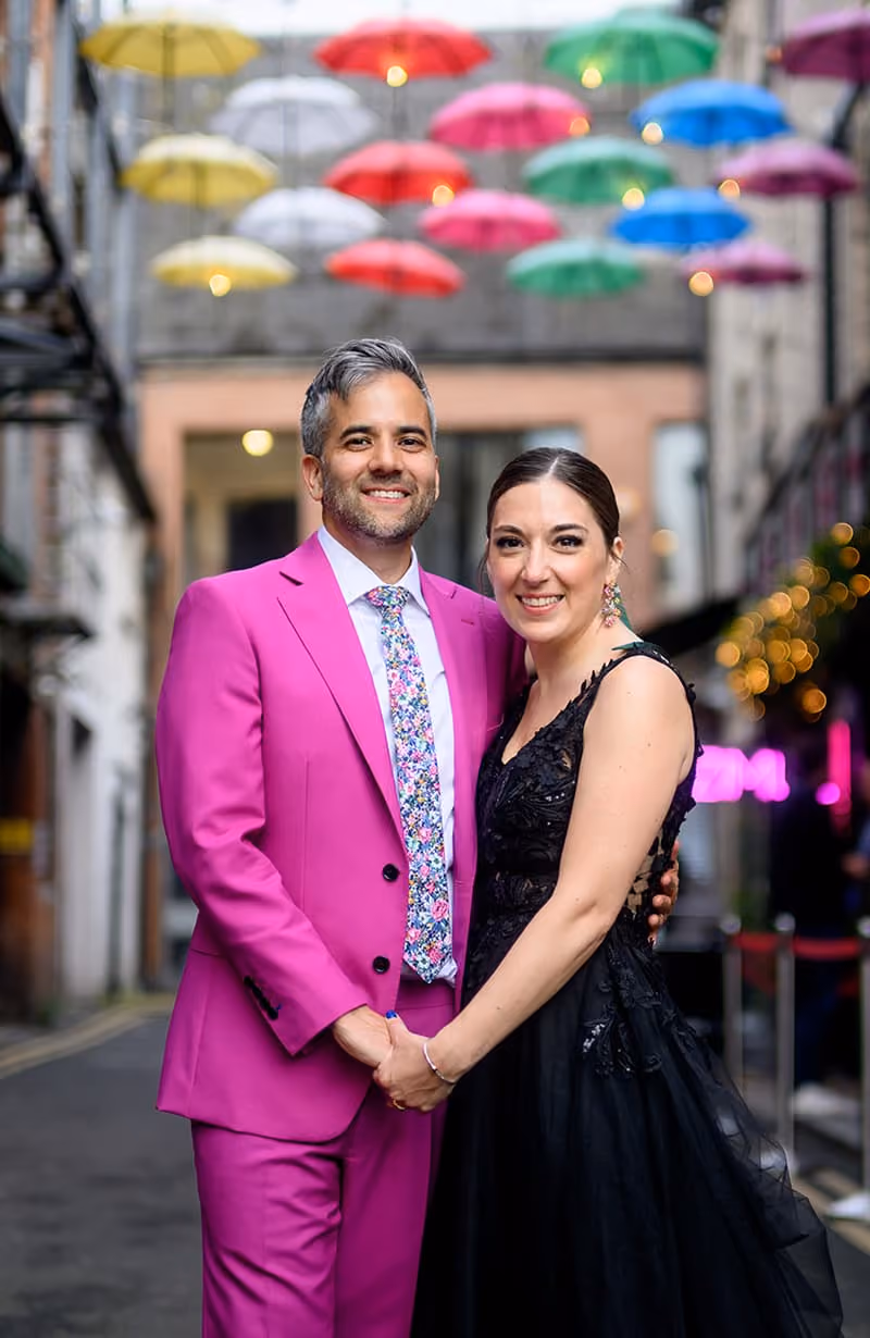 Man in bright pink suit and floral tie holding hands with woman in black dress under colorful umbrellas.