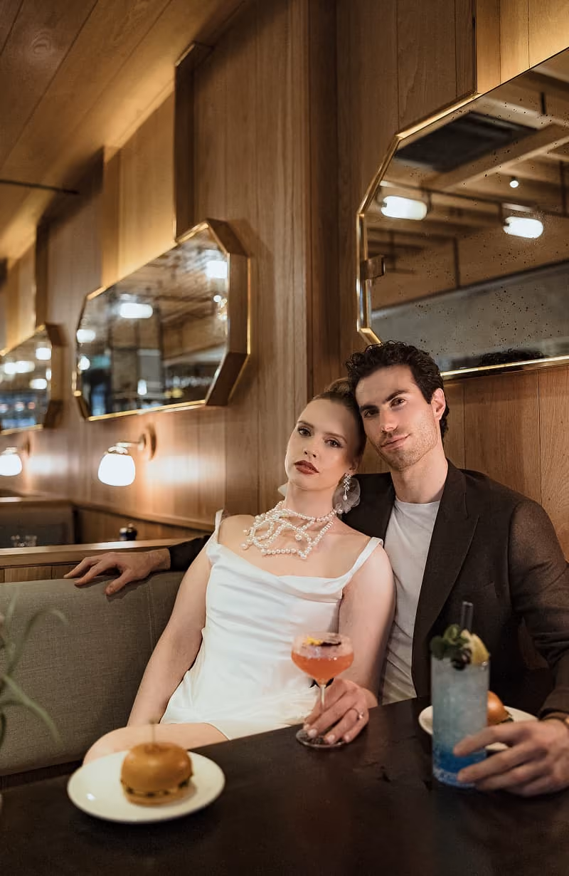 A couple sitting in a booth with a cocktail and burger on the table in a wood-paneled room.