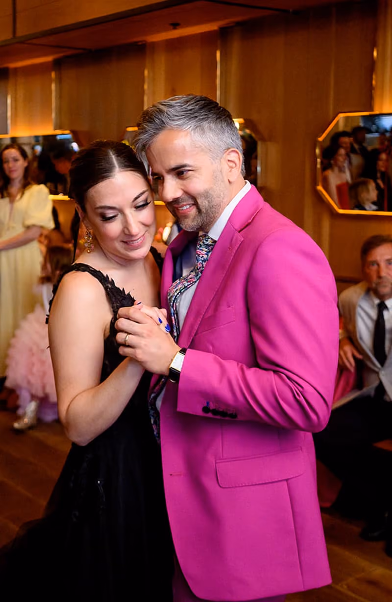 Couple dancing closely at an indoor event, man in pink suit and woman in black dress.
