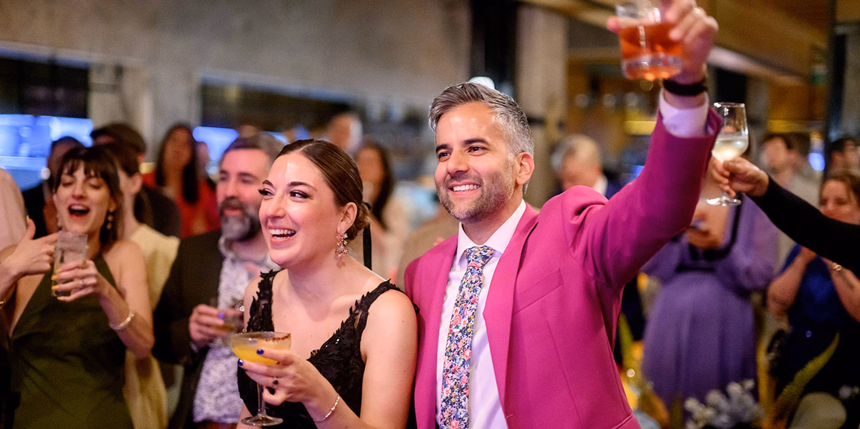 A groom in a bright pink suit raising a glass with guests during a wedding reception.