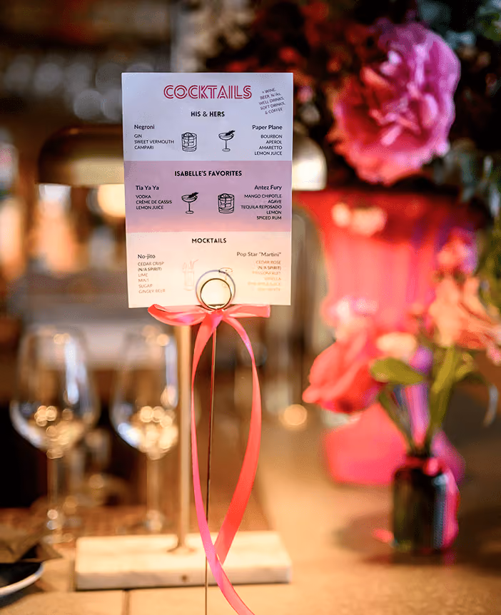 A cocktail menu tied with pink ribbon on a bar, surrounded by flowers and glassware.