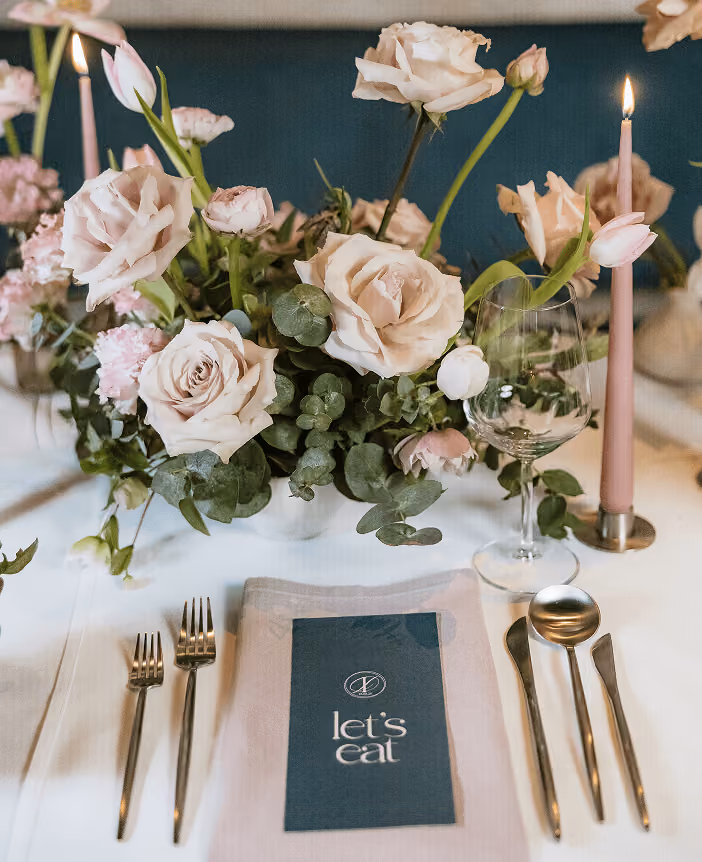 A place setting with blush roses, tapered candles, gold cutlery and a card reading 'let's eat'.