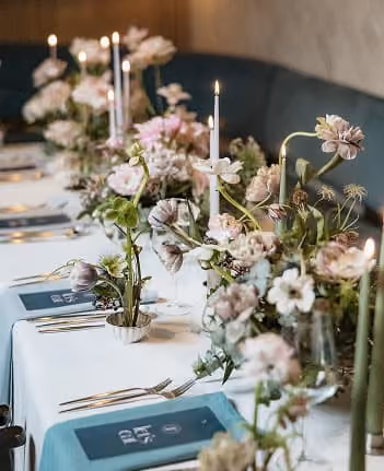 A long wedding table dressed with blush blooms and tall glass stems against a green banquette.