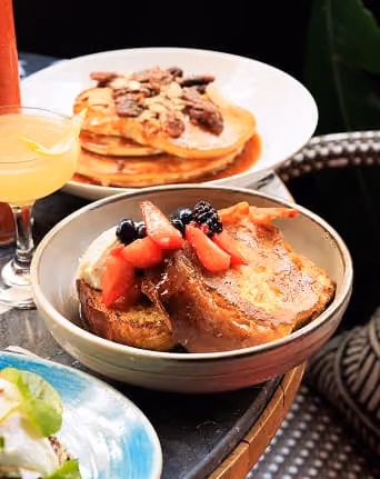 French toast topped with berries and cream in a bowl, with pancakes and a drink behind on a table.