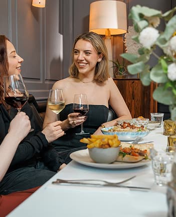 Two women sitting at a restaurant table holding wine glasses and smiling during a meal.