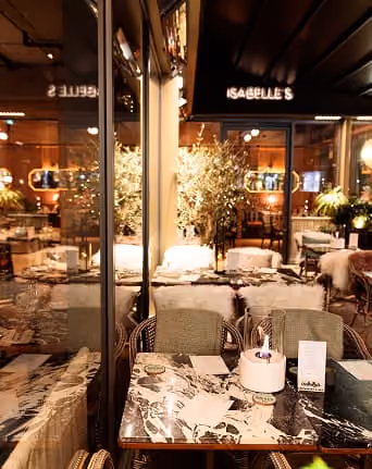 Cozy restaurant table with marble top, candles, menus, and warm indoor lighting with plants in background.