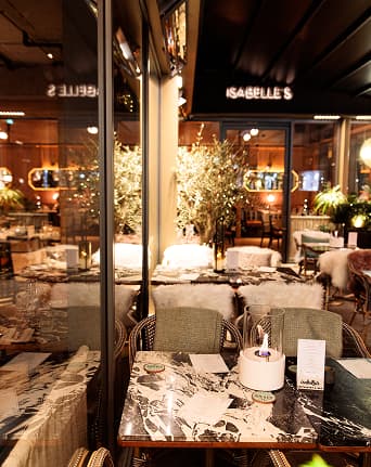 Cozy restaurant table with marble top, candles, menus, and warm indoor lighting with plants in background.