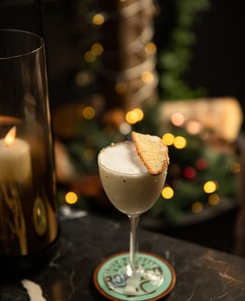 Cocktail glass with creamy white drink garnished with a biscuit, next to a lit candle on a dark table.