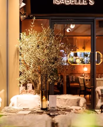 Decorated table with olive branch centerpiece and candles outside a warmly lit restaurant named Isabelle's.