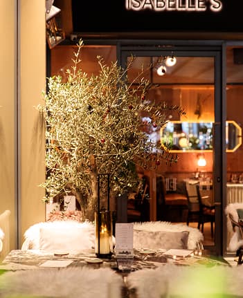 Decorated table with olive branch centerpiece and candles outside a warmly lit restaurant named Isabelle's.