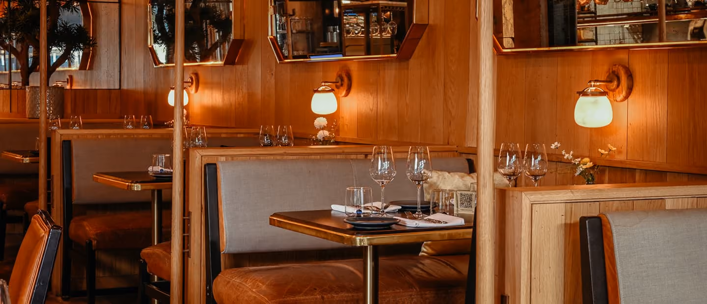 Cozy restaurant booth seating with tan leather cushions and wooden paneling, set with wine glasses, plates, and small flower vases under warm wall lights.