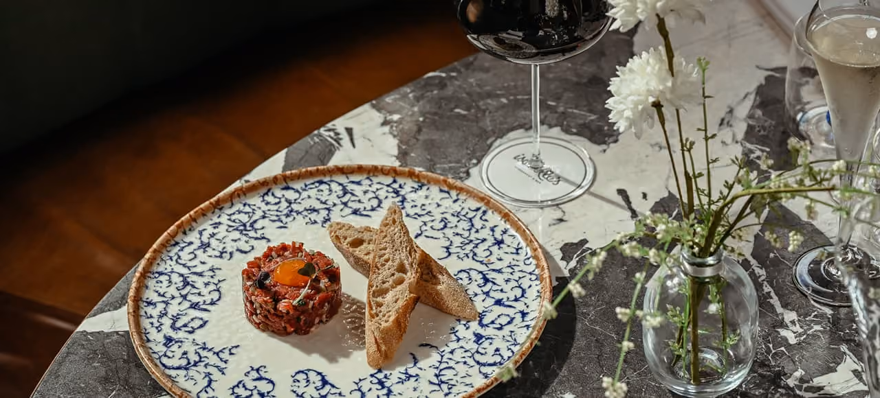 A blue and white patterned plate on a marble table holds two slices of toasted bread next to a round serving of steak tartare topped with a raw egg yolk. Nearby are a glass of red wine, a glass of champagne, and a small vase with white flowers and greenery.