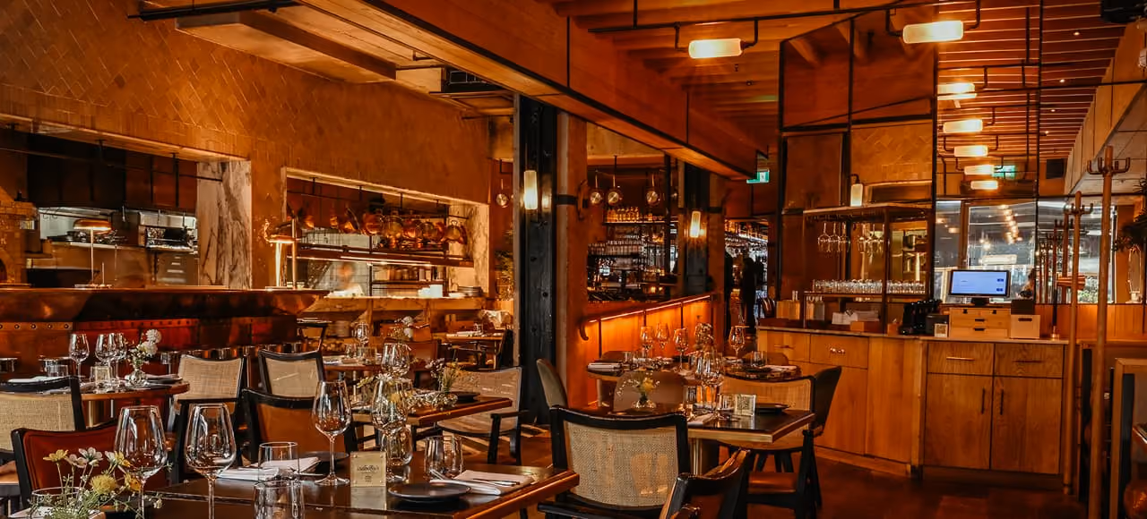 Cozy, warmly lit restaurant interior with wooden beams, tables set with wine glasses, plates, and floral centerpieces, and a wooden service counter with a computer monitor.