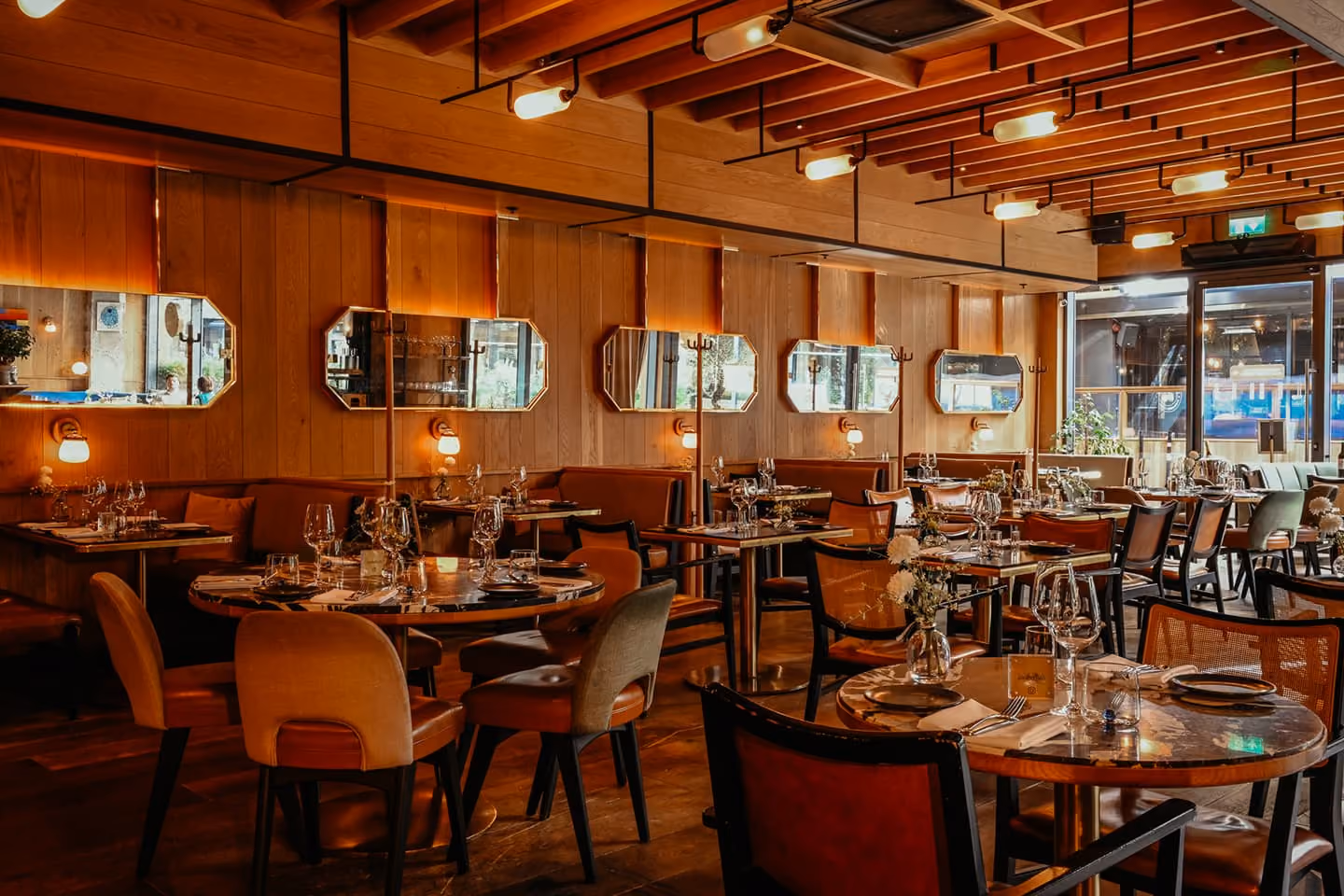 Warmly lit upscale restaurant interior with wooden ceiling and paneling, round and square tables set with glassware, plates, cutlery, and candles, surrounded by cushioned and wooden chairs, mirrors on the walls, and large windows letting in natural light.