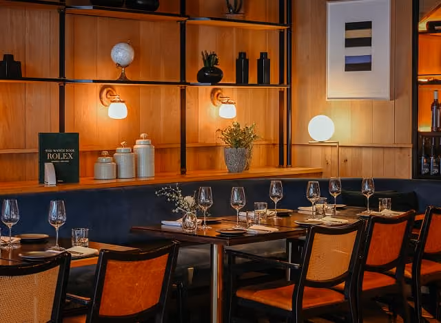 Cozy restaurant interior with wooden panel walls, black cushioned bench seating, wooden chairs, neatly set tables with wine glasses, plates, and small flower vases, warm ambient lighting from wall lamps and a round table lamp, and decorative items like white jars, a globe, and abstract art on the wall shelves.
