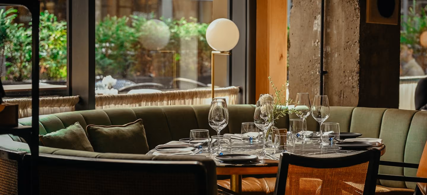 Cozy restaurant table set for four with wine glasses, plates, utensils, a small flower vase, and a round white lamp, surrounded by cushioned green bench seating and wicker chairs, with large windows showing green foliage outside.