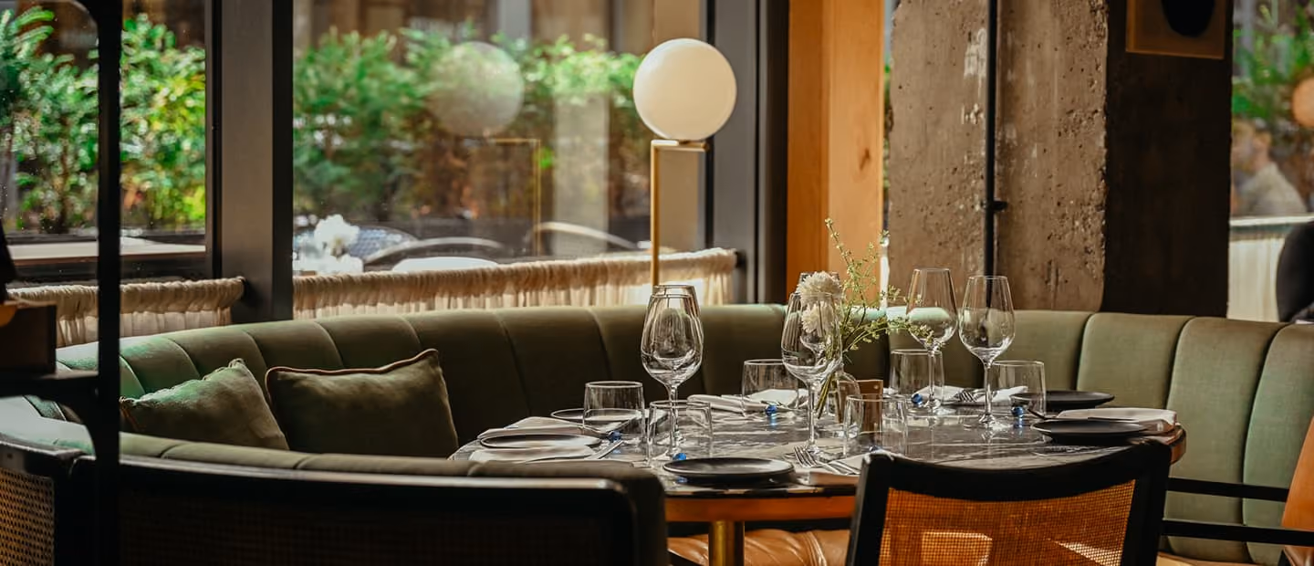Cozy restaurant seating area with a curved green velvet sofa, round wooden table set with plates, wine glasses, cutlery, and a small vase with white flowers, next to a large window with a view of greenery outside.