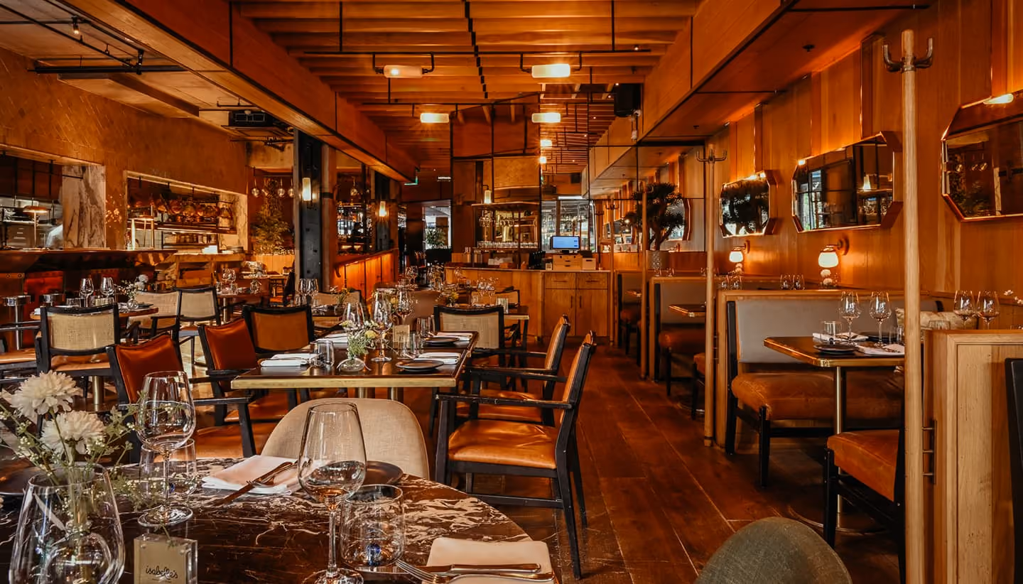 Warmly lit restaurant interior with marble and wooden tables set with wine glasses, plates, cutlery, and floral centerpieces, surrounded by wooden chairs and booths.