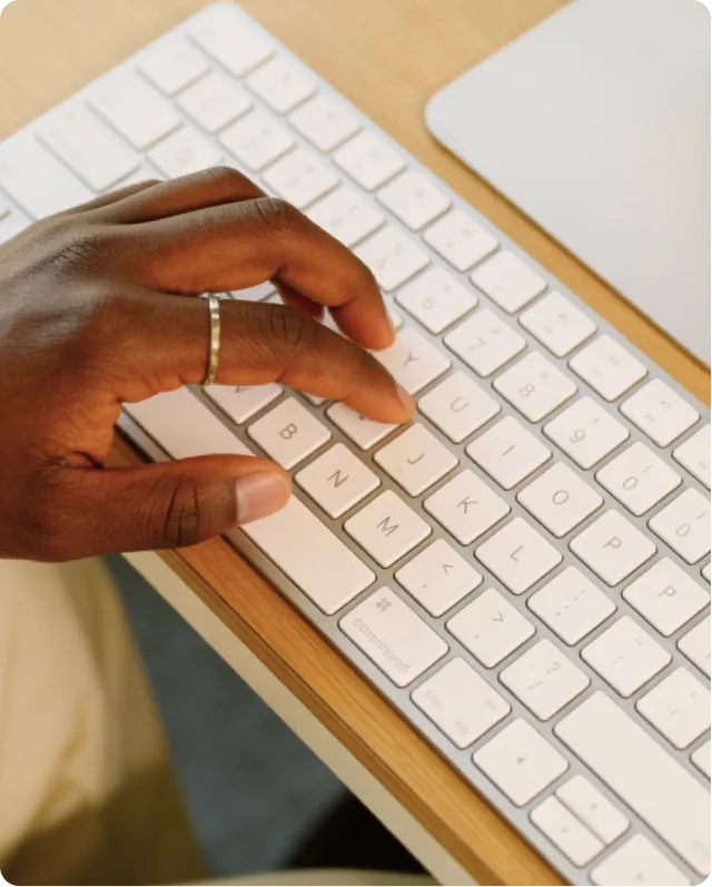 Main noire avec bague tapant sur un clavier blanc posé sur un bureau en bois.