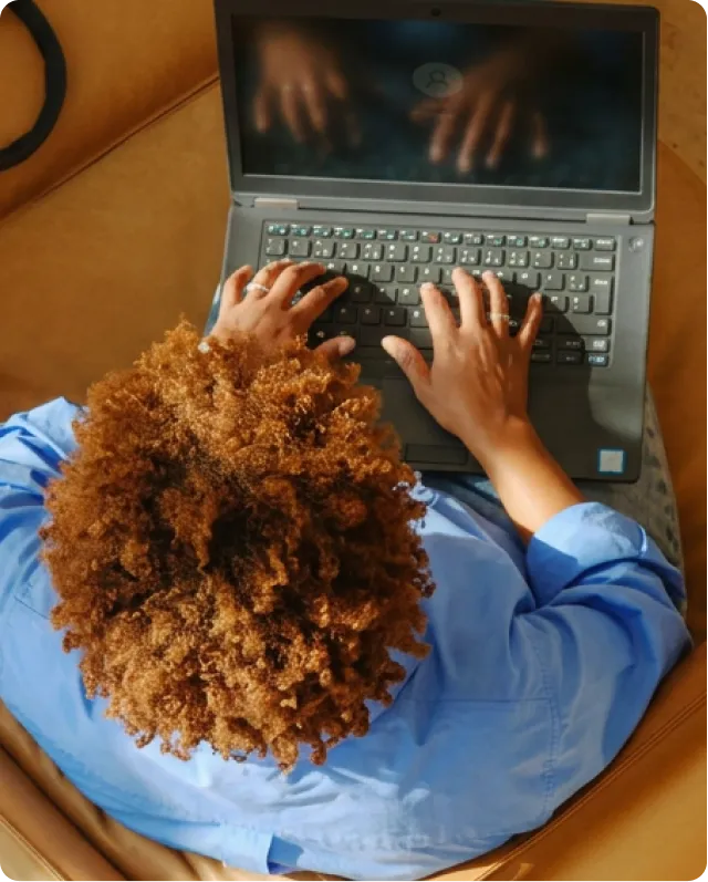 Personne avec cheveux bouclés tape sur un ordinateur portable noir en chemise bleue.