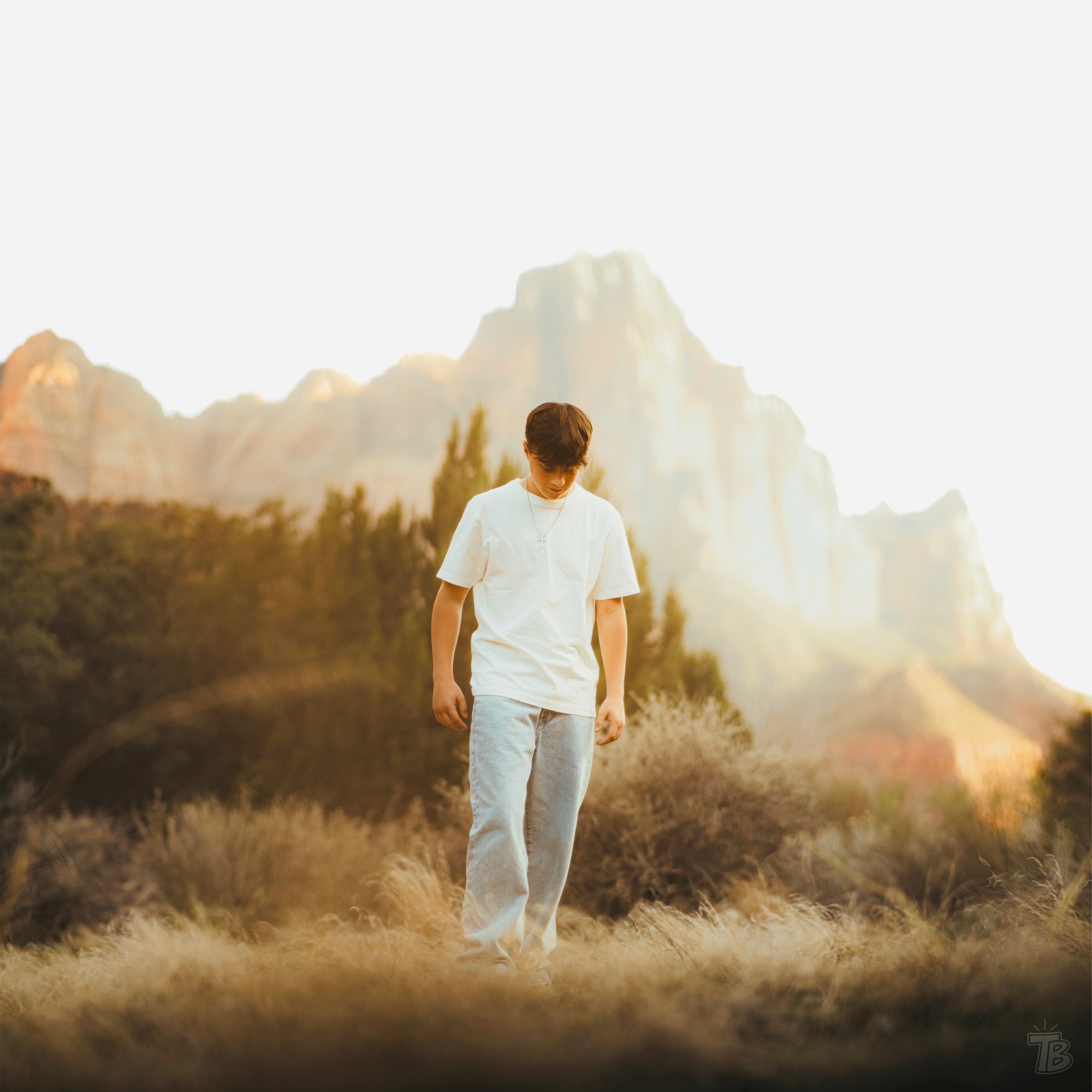 Young man in white shirt and jeans walking in tall dry grass with mountain and trees in background.