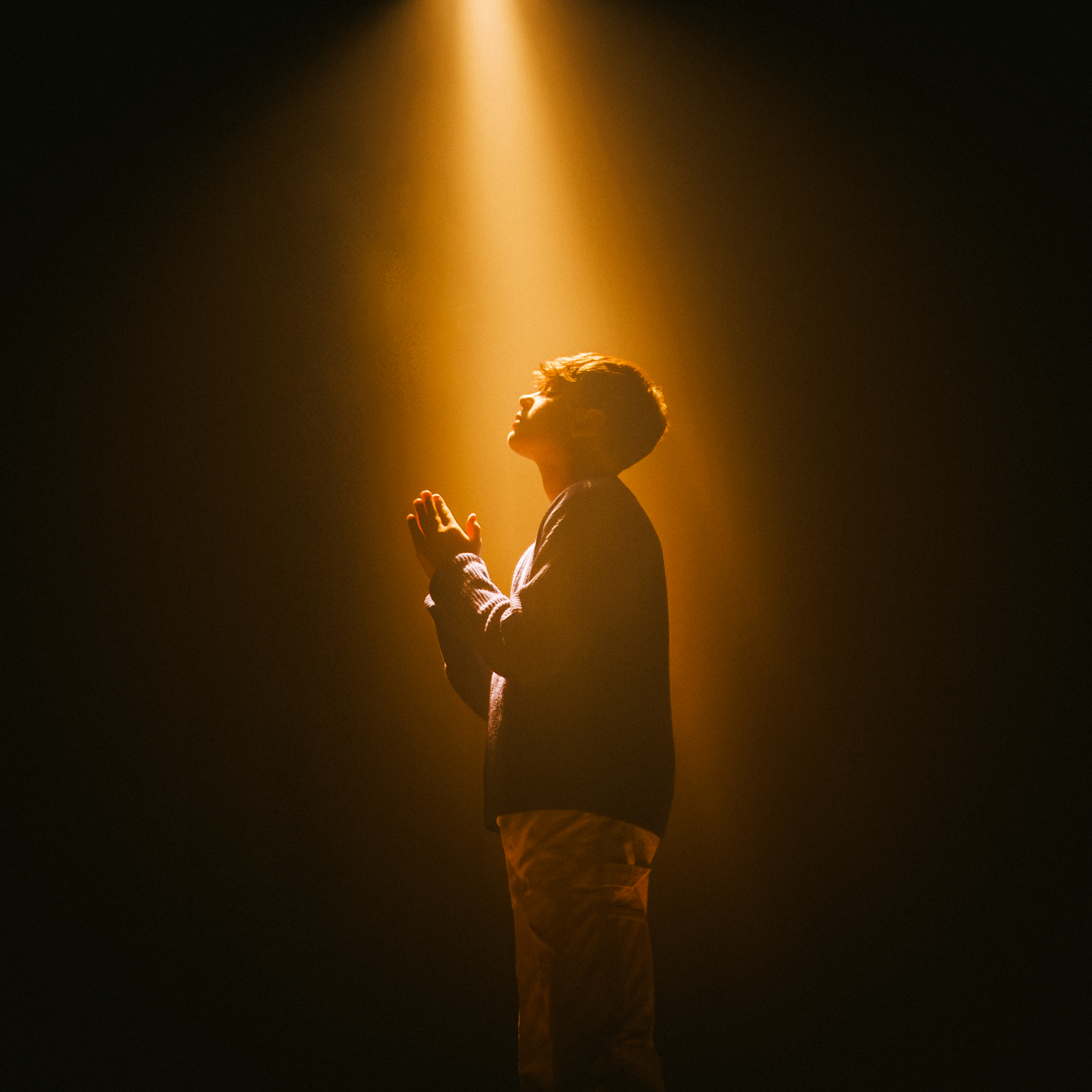Boy standing with hands clasped, looking upward in a warm spotlight against a dark background.