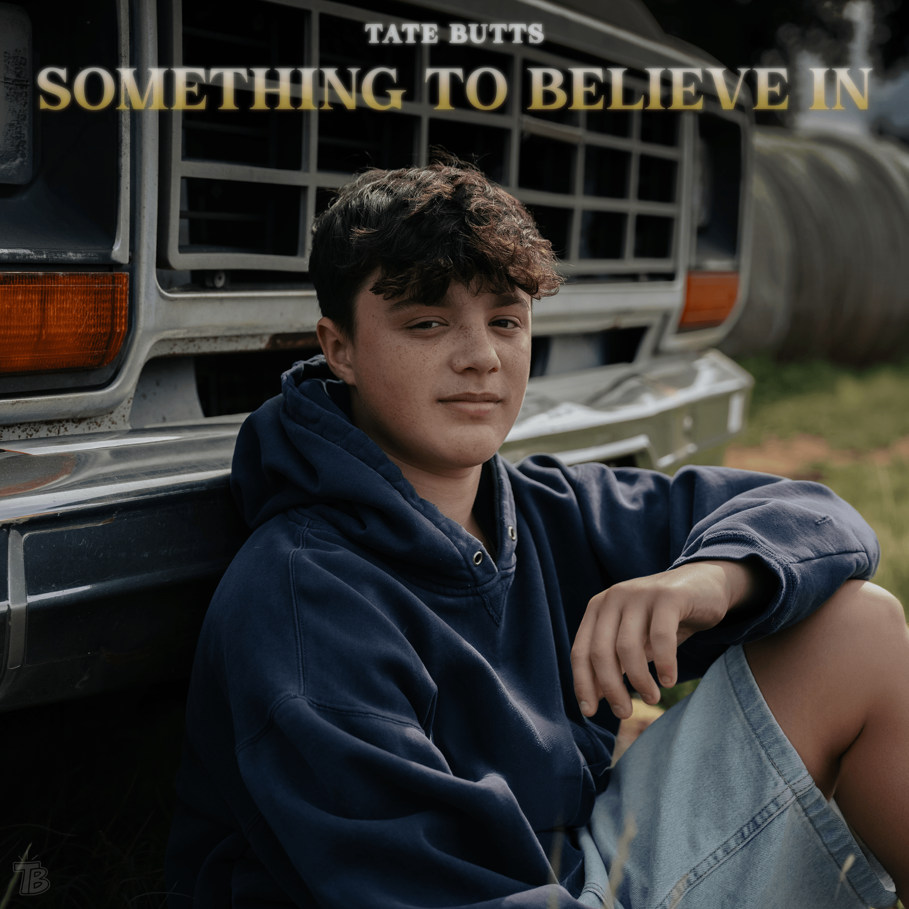 Teen boy in blue hoodie and denim shorts sitting by a truck grill outdoors with a calm expression.