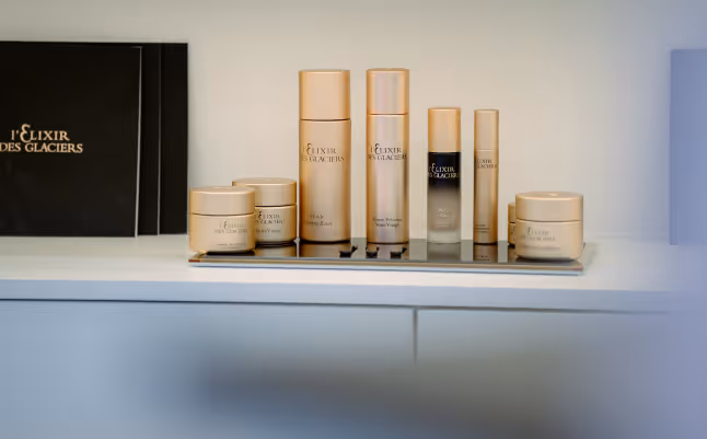 Set of gold-colored skincare products from L'Elixir Des Glaciers arranged on a tray on a white surface.