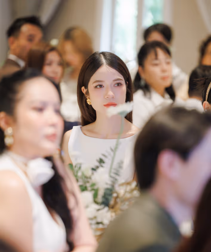 Young woman in white dress holding flowers among a seated audience at an indoor event.