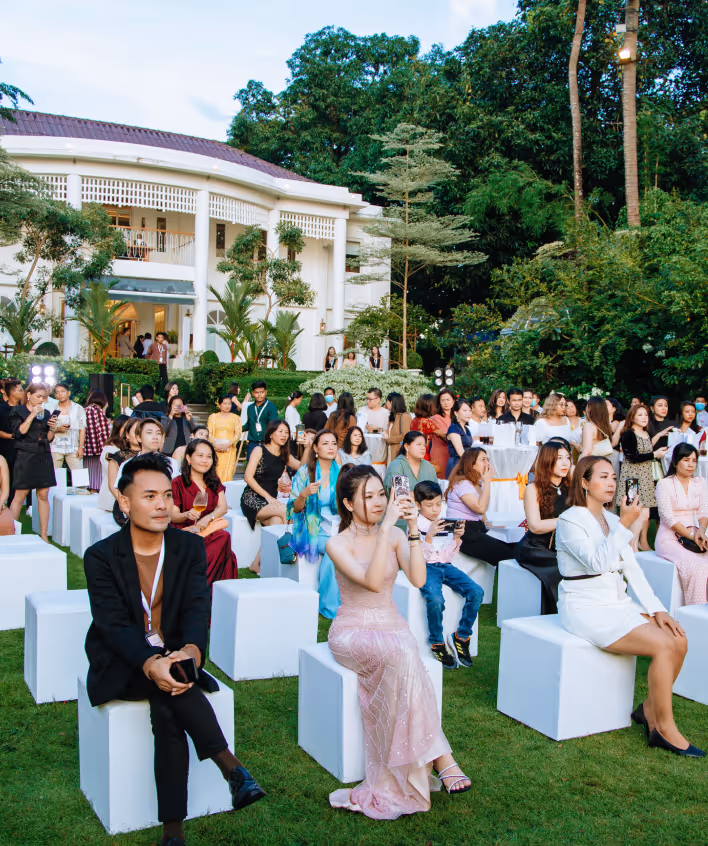 People seated on white cube seats outdoors in front of a large white building during an event.