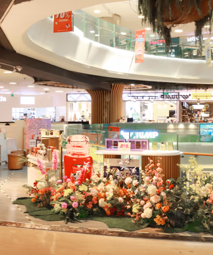Mall interior with a floral display and a large red Yankee Candle jar at a promotional booth.