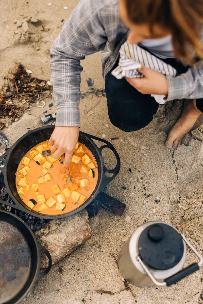 Person stirring a pot of stew with vegetables cooking over a beach campfire on sand.