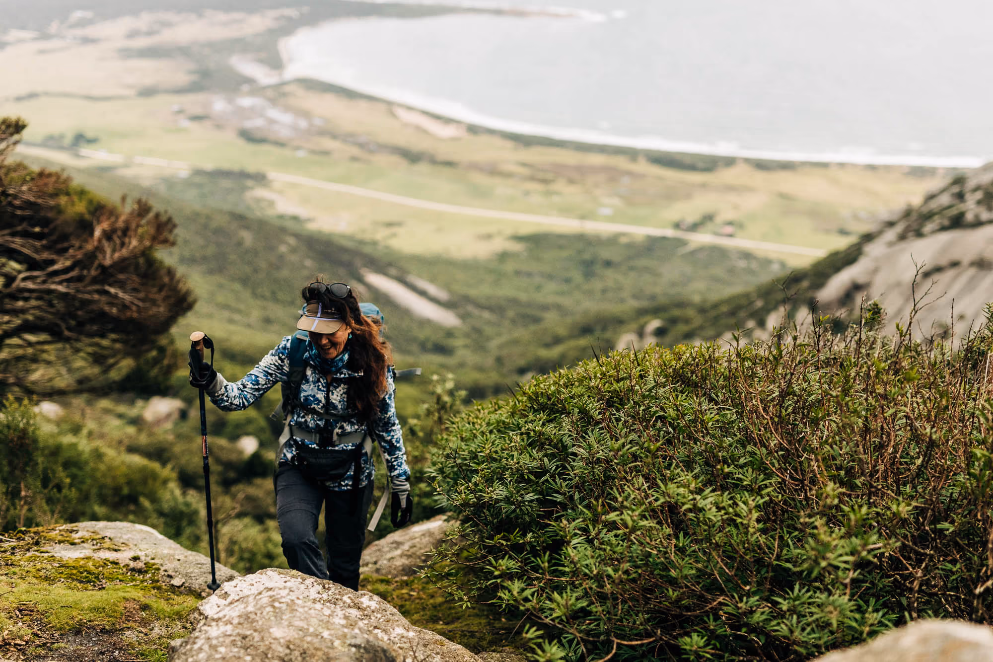 Woman hiking uphill on a rocky trail with trekking pole and backpack, overlooking green valley and sea.