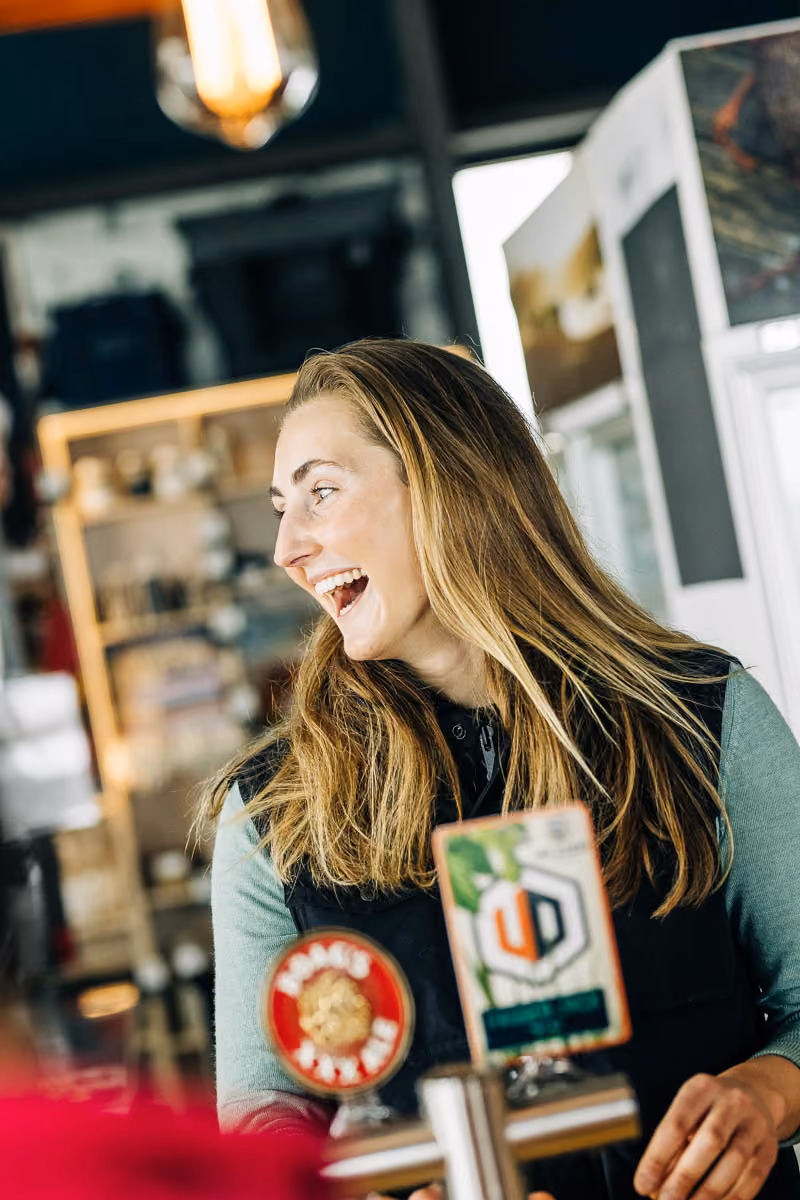 Smiling woman with long hair standing behind a bar counter with beer taps.