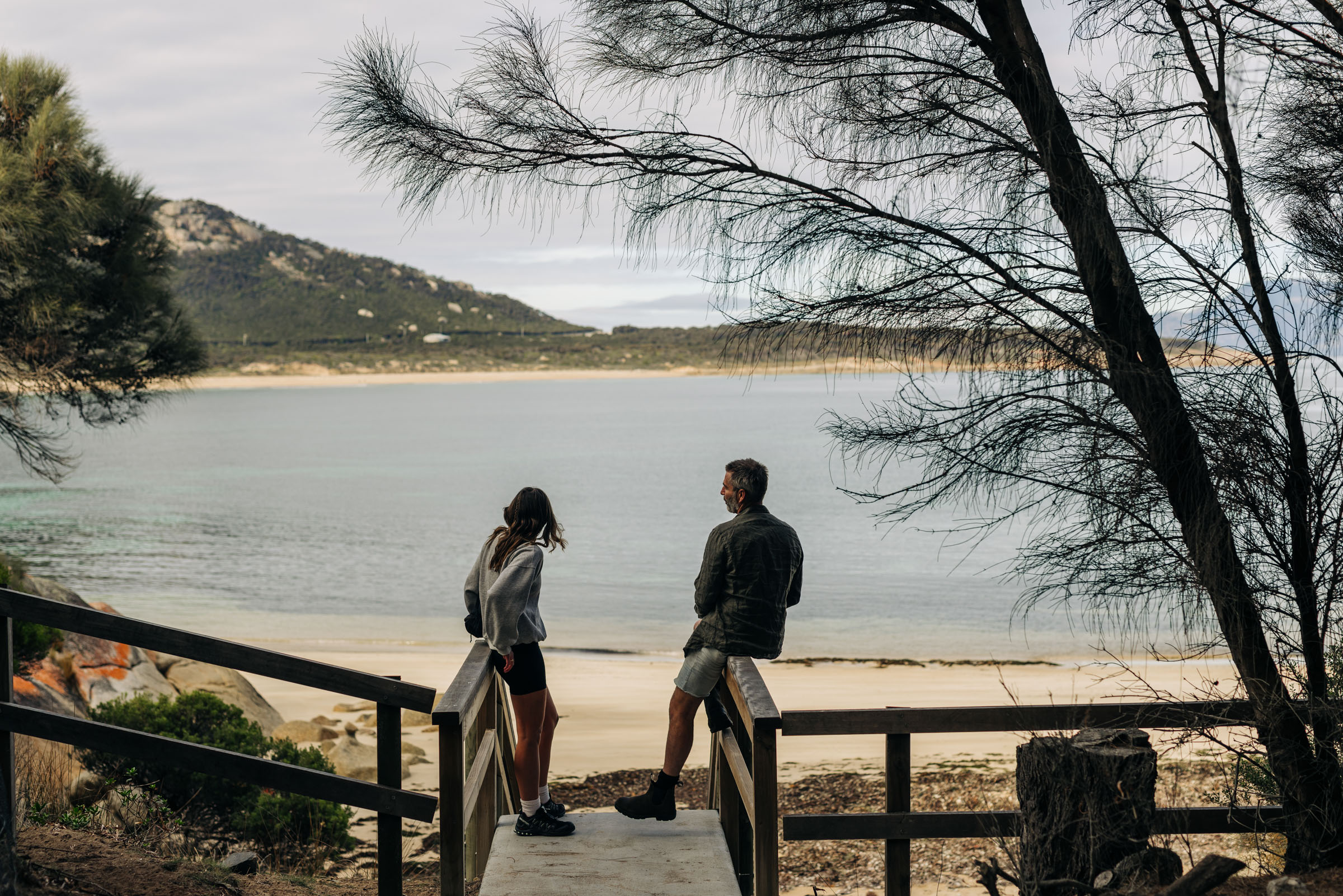 Two people talk on a wooden platform overlooking a calm beach and mountain landscape.