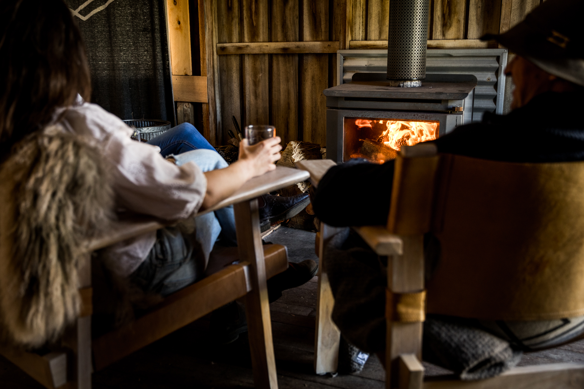 Two people sitting on wooden chairs by a lit wood stove inside a rustic cabin.