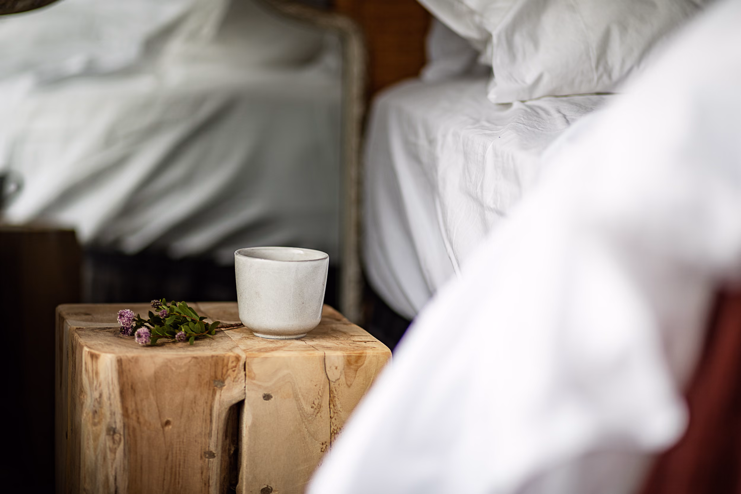 White ceramic cup and small purple flowers on a wooden bedside table next to a white bed.