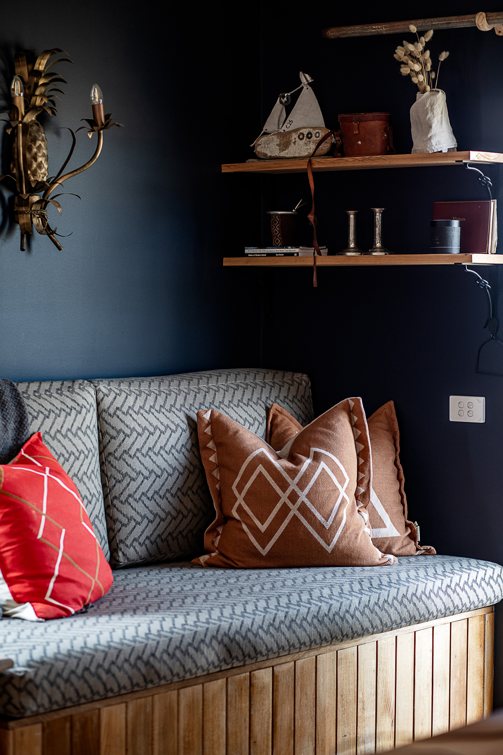 Wooden bench with patterned gray cushions and decorative red and brown pillows under wooden shelves.
