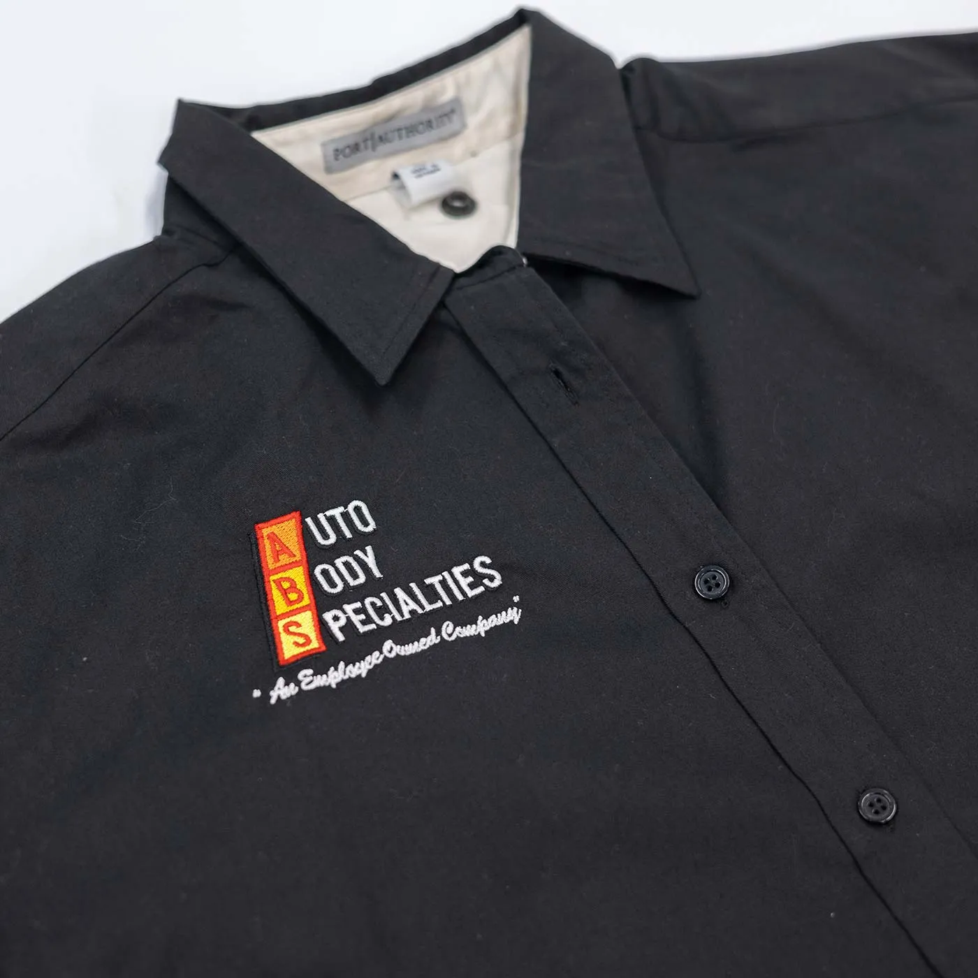 Black collared shirt with embroidered logo on the left chest reading 'ABS Auto Body Specialties' in white and red text, with the phrase 'An Employee Owned Company' beneath it.