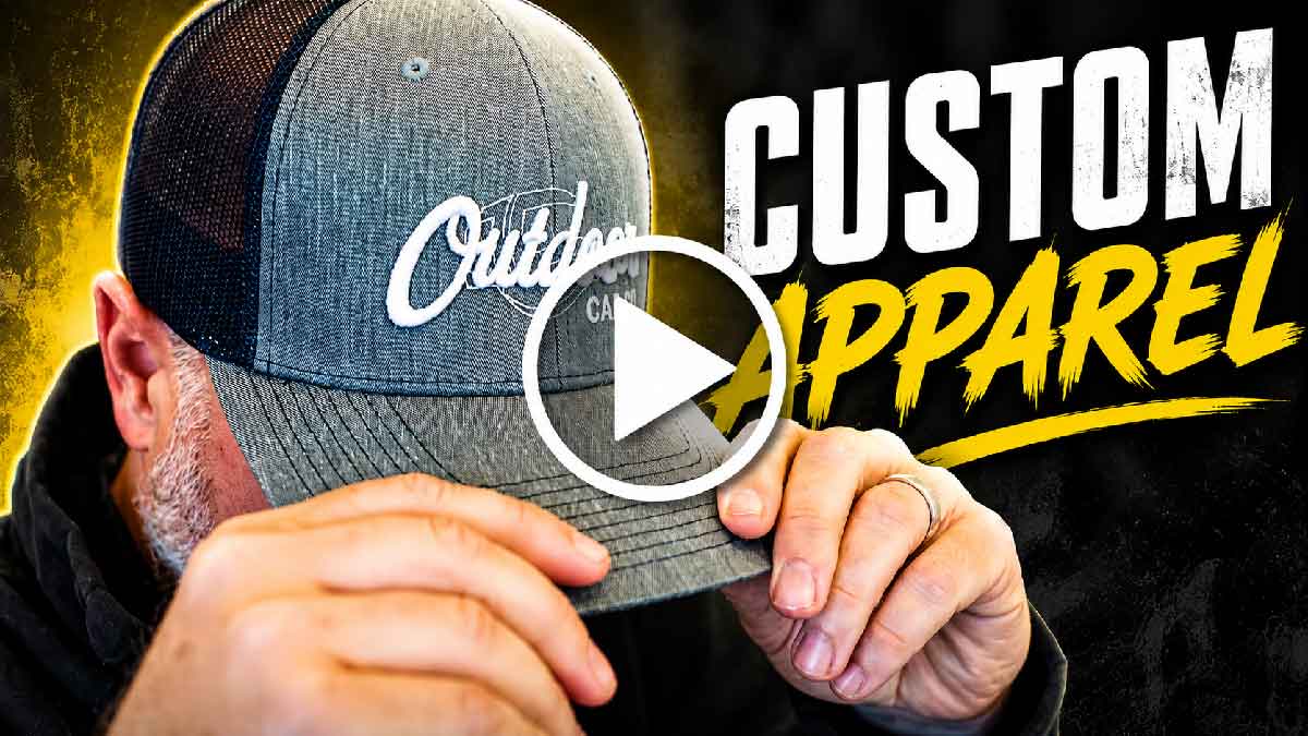 Man adjusting a gray and black mesh baseball cap embroidered with the word 'Outdoor' in white, with the words 'Custom Apparel' in large white and yellow letters on a black background to the right.